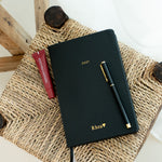 A black 2027 planner with gold foil lettering and a black pen 